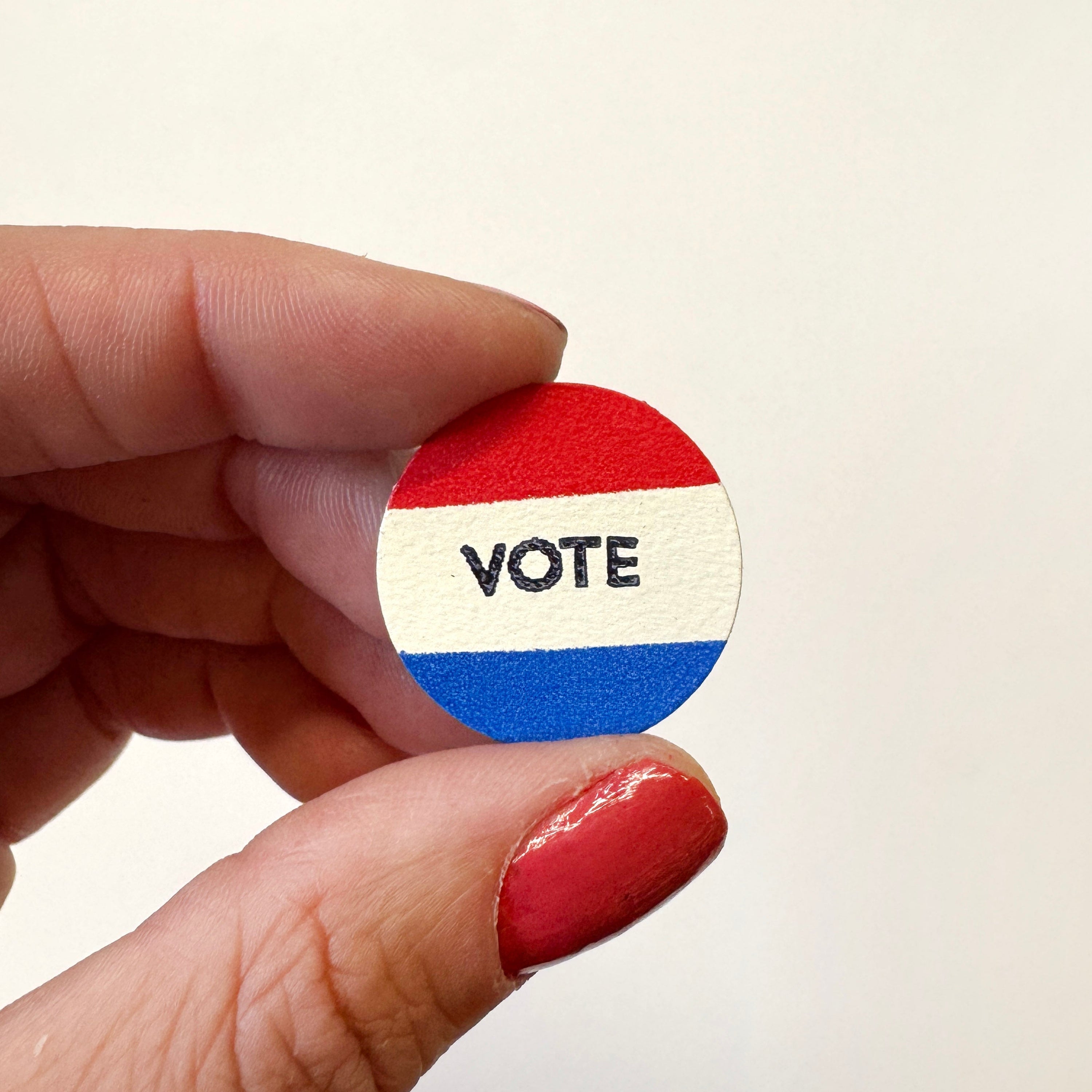 A hand holds a small round magnet featuring red, white, and blue horizontal stripes with the word &