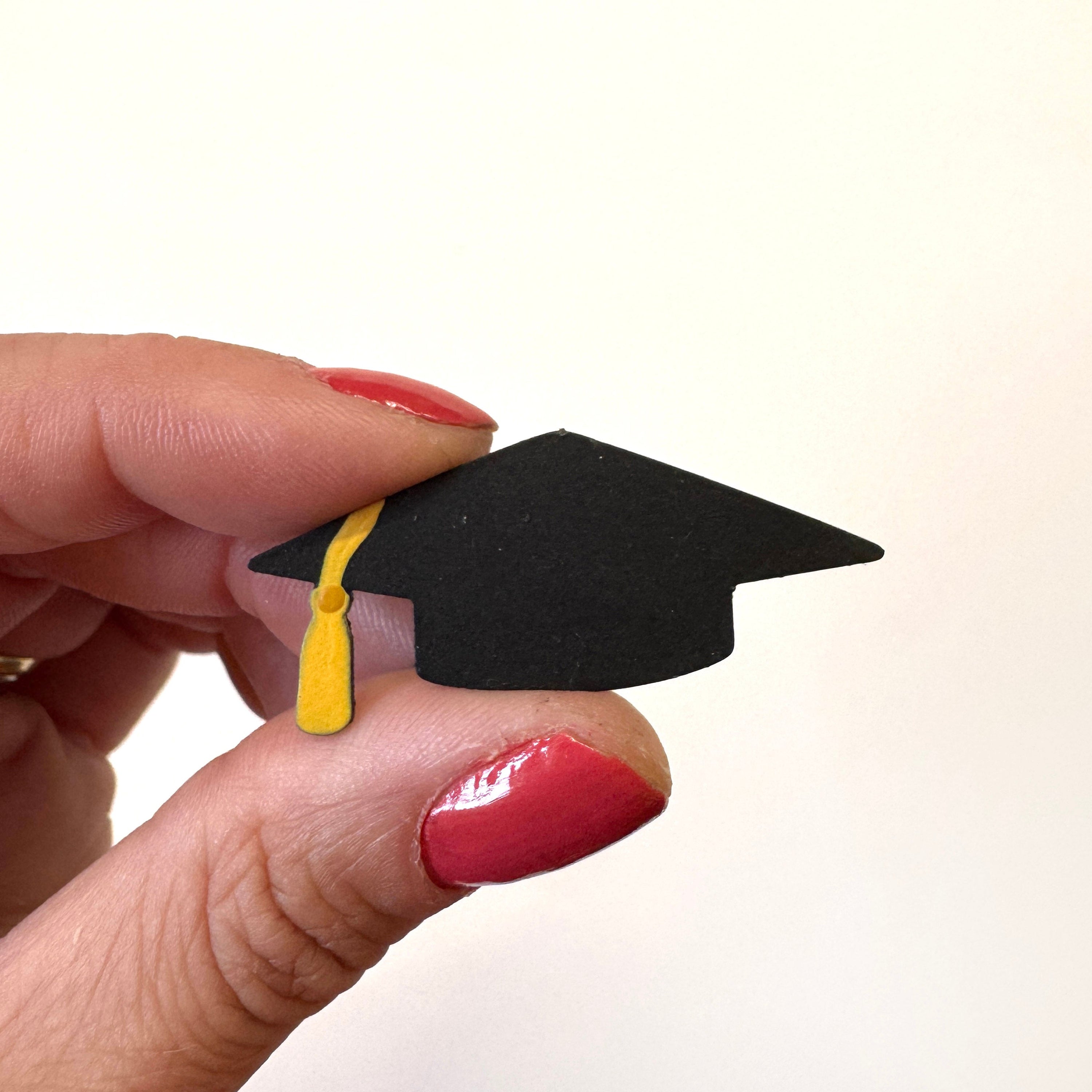 A miniature black graduation cap magnet with a yellow tassel held between fingers against a plain white background.