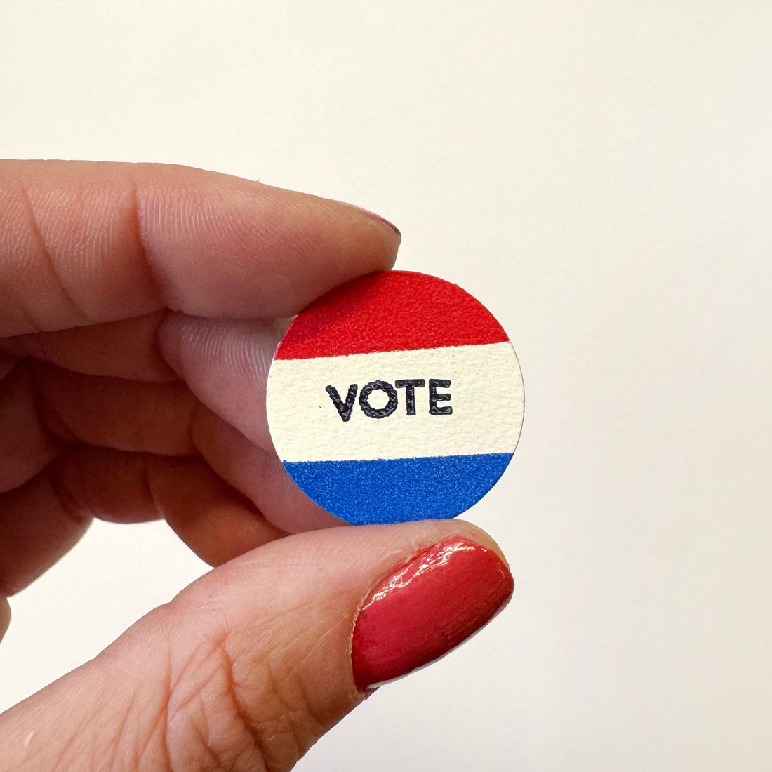 A hand holds a small round magnet featuring red, white, and blue horizontal stripes with the word &