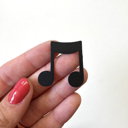 A hand with red nail polish holds a small black music note magnet against a plain white background.