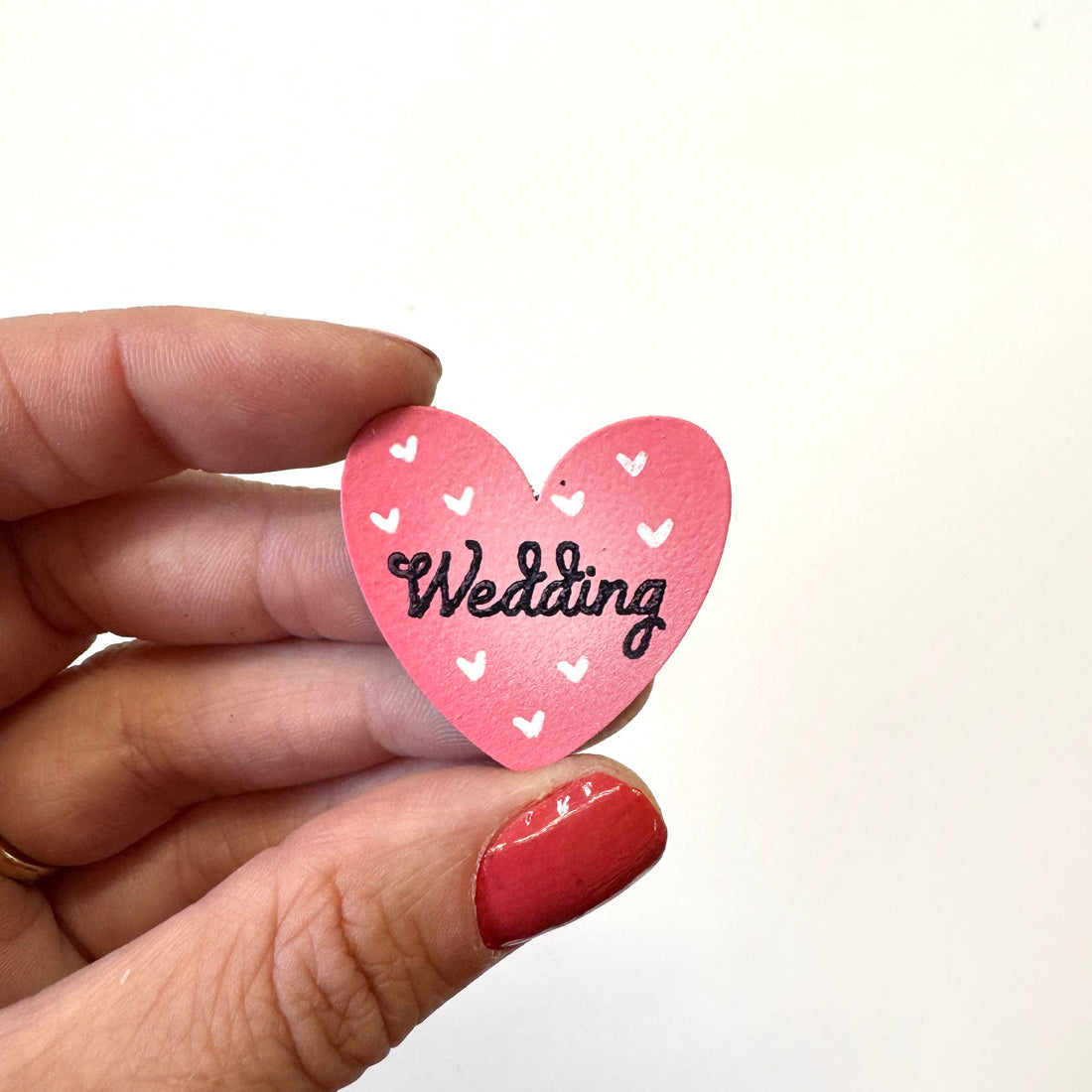 A hand holds a small pink heart-shaped magnet with white heart details and the word &