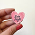 A hand holds a small pink heart-shaped magnet that reads &