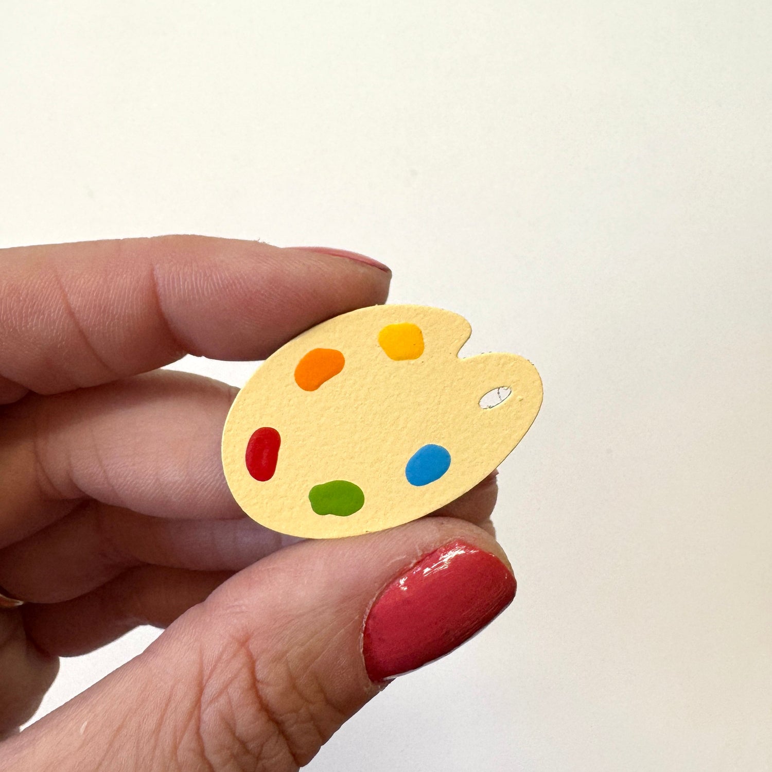 A hand holds a small yellow artist palette magnet with colorful paint dots against a plain off-white background.