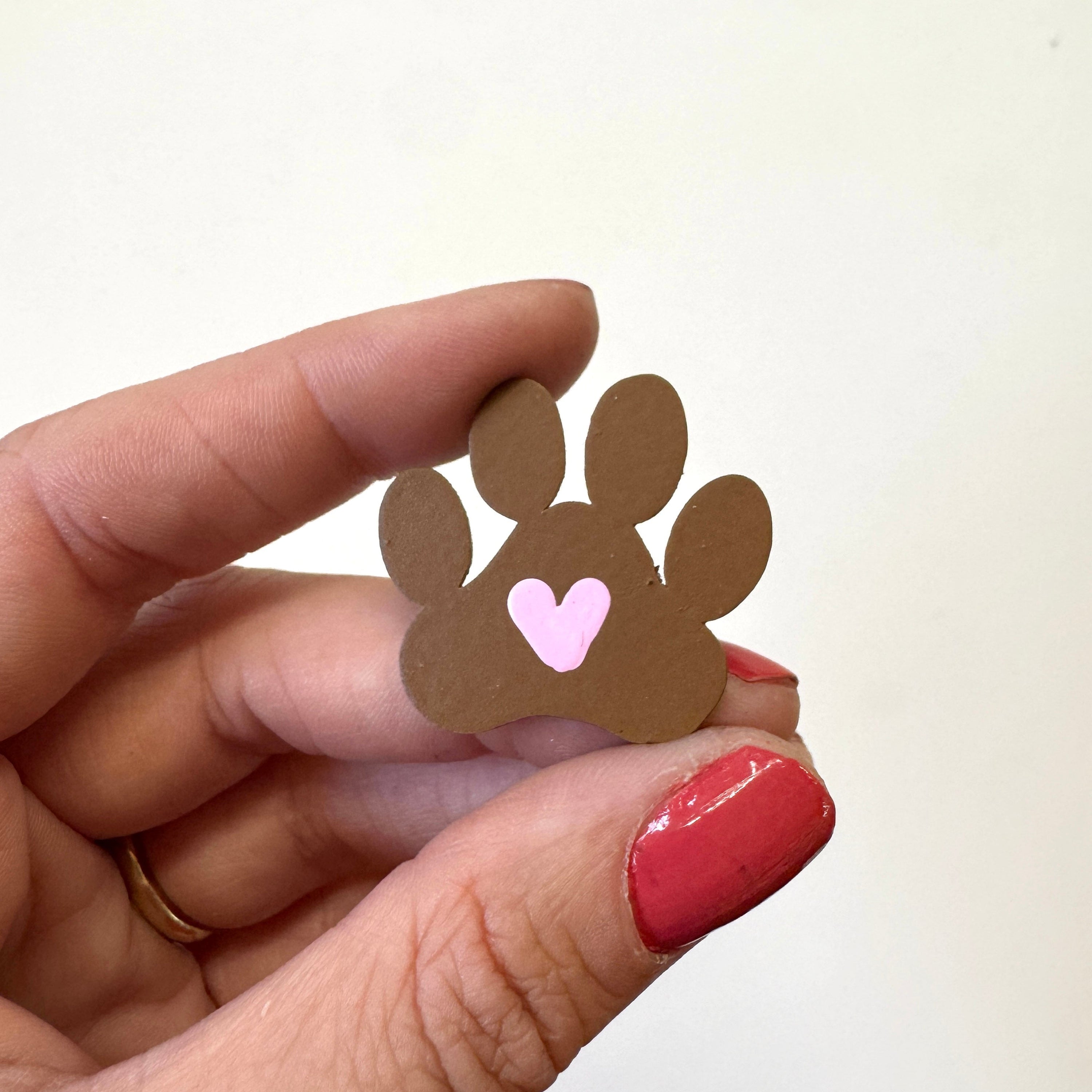A hand with red painted fingernails holds a small brown paw print magnet with a pink heart in the center against a plain off-white background.