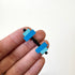 A close-up of a hand holding a small dumbbell-shaped magnet with bright blue and light blue rectangular weights on a black handle, against a plain white background.