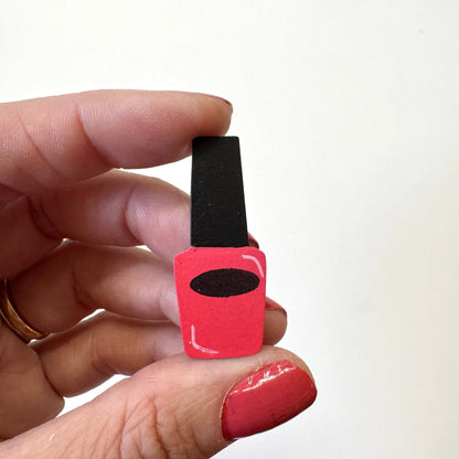 A hand holding a small, hand-painted nail polish bottle magnet. The bottle is painted bright pink with a black cap and a black oval detail on the front, set against a plain white background.