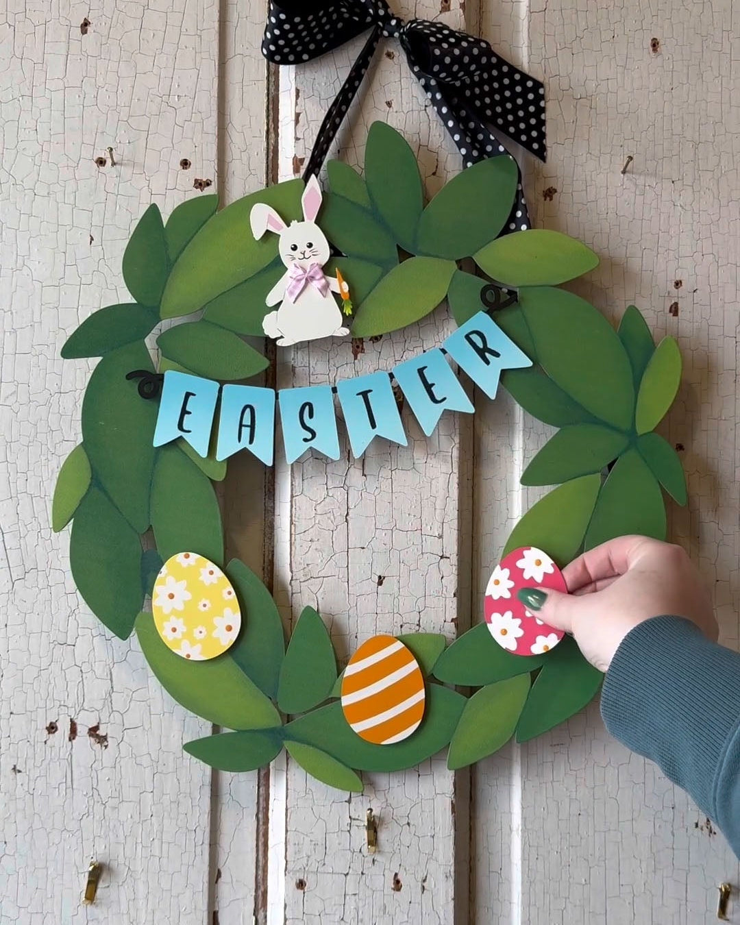 A green leaf wreath with Easter decorations hanging on a distressed white wooden door. The wreath features a black polka-dot bow, a blue &